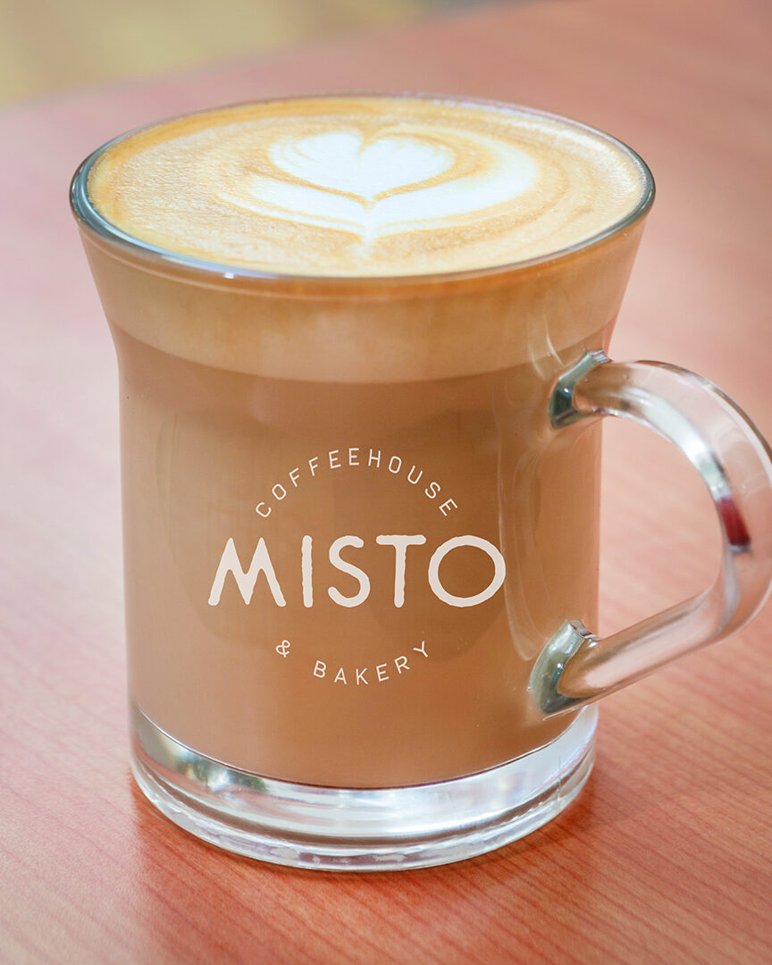 A glass Misto mug of latte with heart latte art on a wooden table.