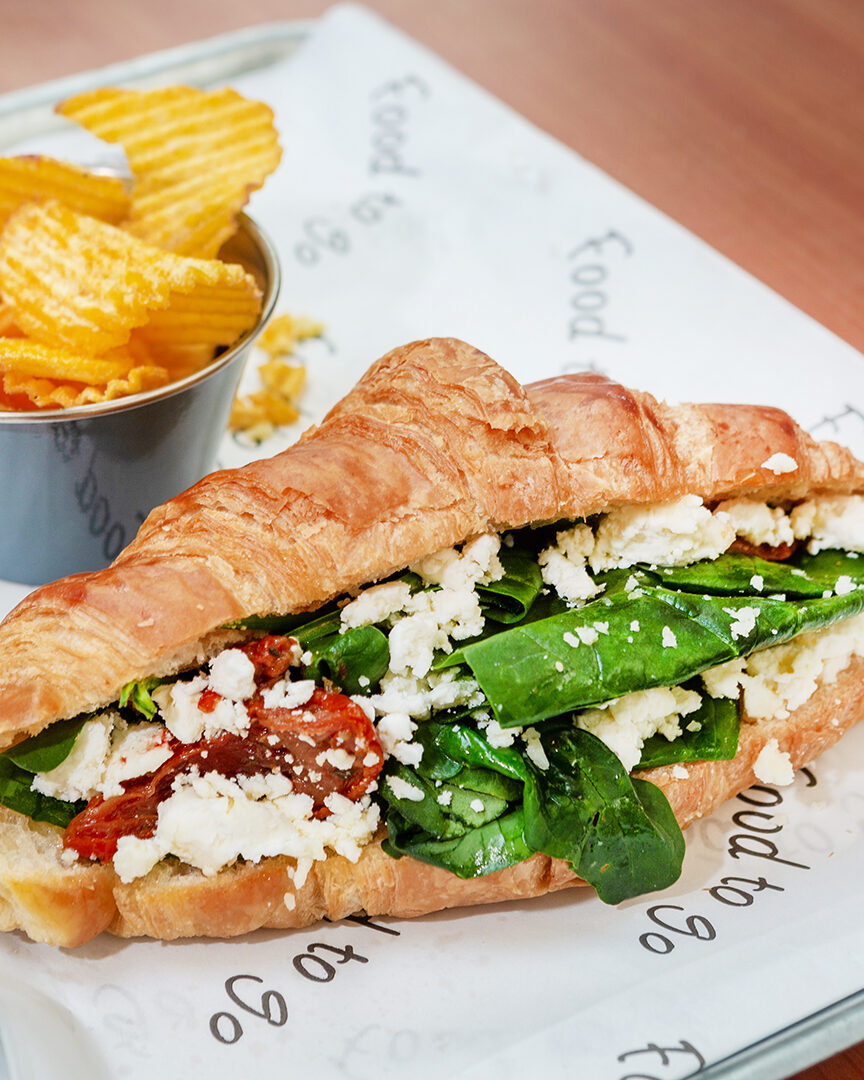 A filled croissant with spinach, feta and sun-dried tomato, with a pot of ridged crisps, on a paper-lined tray.